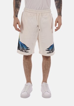 Mand iført off-white snøreshorts med blå og sorte fuglevinge-print nær kanten, kombineret med hvide sneakers og en hvid skjorte.