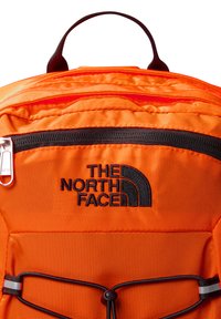 Pomarańczowy plecak z materiału z czarnym zamkiem błyskawicznym i czarnym haftowanym logo. Posiada szczegóły z gumką oraz uchwyt do noszenia na górze. Gładka faktura.