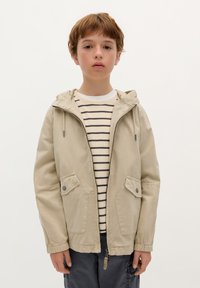 Giacca leggera beige con cappuccio, chiusura a zip, due tasche frontali e polsini elasticizzati, indossata sopra una camicia a maniche lunghe a righe.