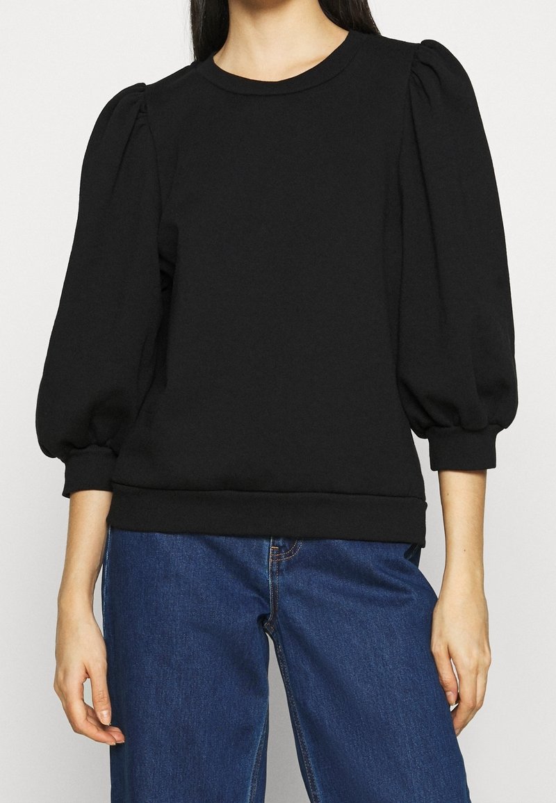 Femme portant un haut noir à manches bouffantes trois-quarts et un jean en denim bleu, debout devant un fond uni.