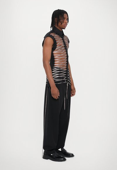Chemise sans manches noire avec des panneaux transparents et des détails en lacets argentés ; associée à un pantalon noir ample et des chaussures noires ; design minimaliste.