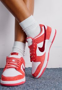 Červené a biele tenisky Nike s povrchom z kože a semišu, ktoré majú výrazné logo swoosh a šnúrky, spárované s bielymi ponožkami po členky.