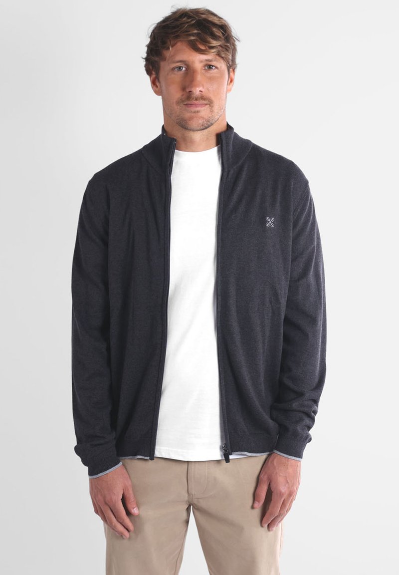 Veste zippée gris foncé avec col montant, manches longues et poignets élastiques, superposée à un t-shirt blanc. Tissu lisse avec une texture subtile.