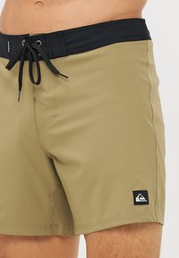 Khaki plavky s čiernym elastickým pásom, šnúrkou a bočnými vreckami. Obsahuje nášivku s logom na dolnej ľavej nohe. Hladká textúra.
