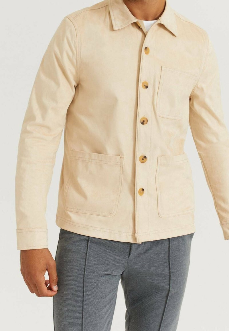 Homme portant une veste beige boutonnée avec trois poches avant et un pantalon gris, debout devant un fond uni.