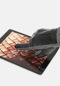 Juodos odinės ir pilkos mezginio pirštinės liečia planšetinio kompiuterio ekraną, kuriame rodoma tekstūrinė raštas šiltomis spalvomis, pabrėžiant funkcionalumą skaitmeniniam sąveikai.