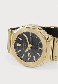 G-Shock käekell kuldse roostevaba terase korpuse, musta tahhiga ja helendavate märkidega. Omab kaht erinevat ajavööndit ja kummiseid aktsente rihmal.