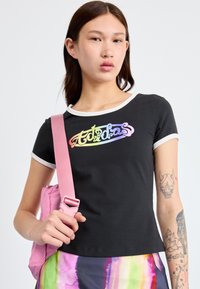 Sort kortærmet T-shirt med regnbuefarvet "Adidas" logo, hvid krave og ærme trim. Modellen holder en lyserød taske og bærer en farverig mønstret nederdel.