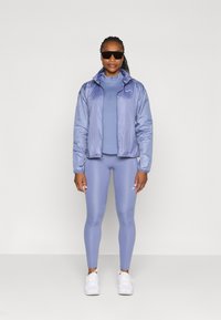 Lys lilla vindjakke med lynlås og hætte, lagd over en matchende top og leggings. Akkompagneret af hvide sneakers og solbriller.