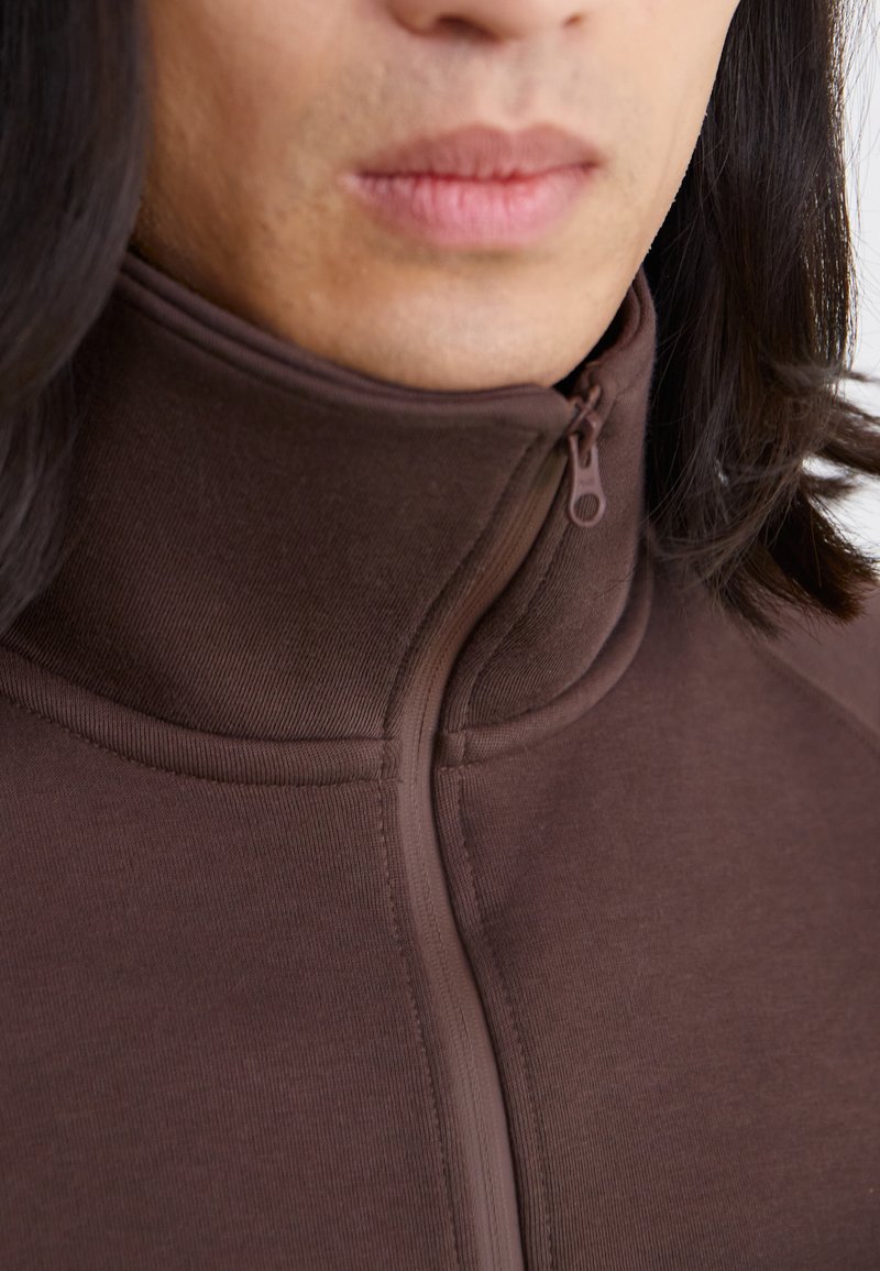 Sudadera marrón con cremallera y cuello alto, fabricada en un tejido suave y con costuras tonales. Cremallera situada en el centro del cuello.
