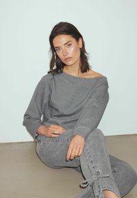 Grijze sweatshirt met brede hals en lange mouwen, gecombineerd met grijze denim broek met strikdetails aan de manchetten. De stof is glad overal.