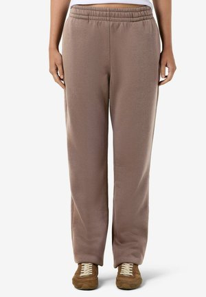 Personne portant un pantalon de survêtement marron ample avec une ceinture élastique et des baskets en daim marron avec des lacets blancs, debout sur fond blanc.