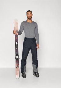 Skikleding met een grijze longsleeve top en zwarte broeken met grijze accenten, houdend een meerkleurige ski met zwarte bindingen.