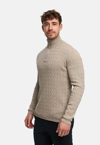 Beige strikkegenser med høy krage, teksturert mønster og halv glidelås foran. Har lange ermer og en tettsittende design.