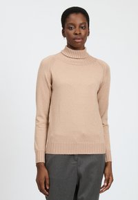 Maglione beige a collo alto con bordo e fondo a coste, tessuto liscio, design aderente e maniche lunghe, indossato sopra pantaloni grigi.