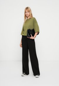 Oversized t-skjorte i olivengrønt, svarte vidbukser, holder en svart clutchveske. Joggesko med hvit såle. Enkel, minimalistisk design.