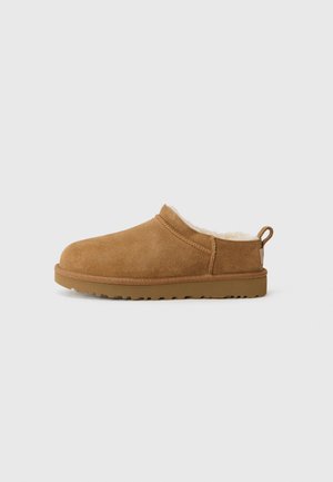 Chausson en daim marron à enfiler avec doublure en polaire blanche et semelle en caoutchouc, vu de profil sur un fond clair uni.