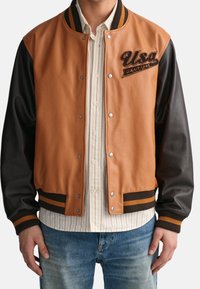 Hombre vistiendo una chaqueta de varsity marrón y negra con un parche que dice "USA GANT 1949", sobre una camisa de botones a rayas beige y jeans azules.