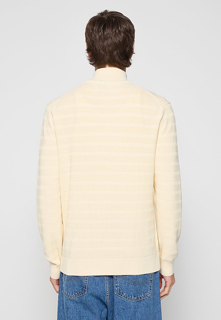 Personne avec des cheveux bruns courts portant un pull à manches longues côtelé couleur crème et un jean en denim bleu, vu de dos.