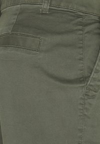 Donkergroen olijfgroen stof met een gladde textuur, voorzien van een achterzak en subtiele stikdetails. Close-up van het materiaal.