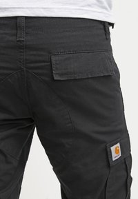 Zbliżenie czarnych spodni roboczych Carhartt, pokazujące tylną kieszeń z klapą oraz wzmocnione szwy w okolicy uda.