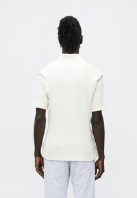 Polo de manga curta branca com textura canelada suave, com colarinho clássico e bainha reta, vista por trás.