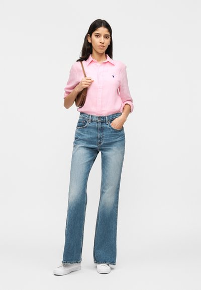 Femme vêtue d'une chemise rose clair boutonnée, jean bleu, baskets blanches et tenant un sac à bandoulière marron, debout devant un fond uni.