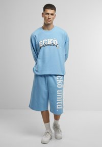 Felpa di cotone azzurro chiaro con collo rotondo, che presenta la scritta "ECKO unlimited" in stampa bianca e nera. Abbinata a pantaloni corti blu coordinati e sneakers bianche.