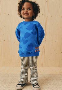Helles blaues Sweatshirt mit floralem Muster und "HAPPY & KIND" in orangem Text; kombiniert mit gestreiften, schwarz-weißen Schlaghosen und Sneakers.