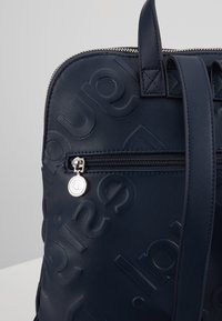 Sac à dos bleu marine avec un design texturé, doté d'une poche zippée à l'avant et de détails en métal argenté. Le tirette de la fermeture éclair comporte un accent en forme de logo circulaire.