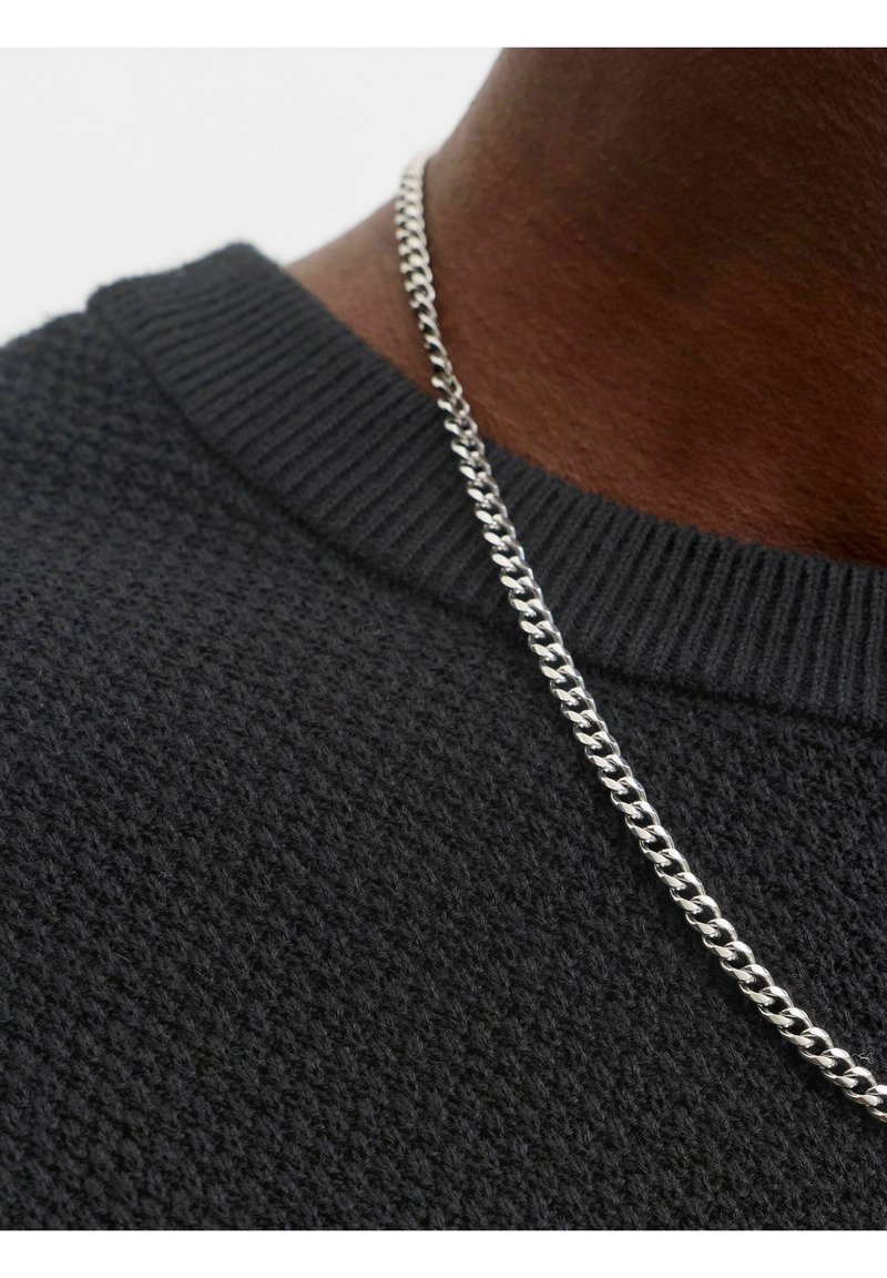 Gros plan d'une personne portant un collier chaîne argenté et un pull en maille noire texturé avec un col côtelé.