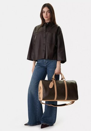 Donna in piedi con capelli lunghi e lisci, indossa una giacca di pelle marrone scuro, jeans a gamba larga blu, tiene una borsa da viaggio marrone con motivo.