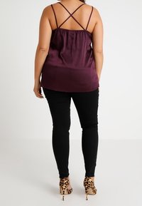 Kvinna i en lila topp med korsade remmar, svarta tajta byxor och högklackade skor med leopardmönster, stående med ryggen mot kameran på en vit bakgrund.