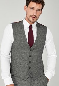 Grå uldvest med en skræddersyet pasform, udstyret med et notched revers, seks knapper og sidelommer. Båret over en hvid skjorte og en granatfarvet slips.