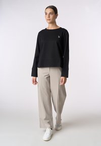Felpa nera a maniche lunghe con scollatura rotonda, caratterizzata da un piccolo emblema, abbinata a pantaloni beige chiaro a gamba larga e sneaker bianche.