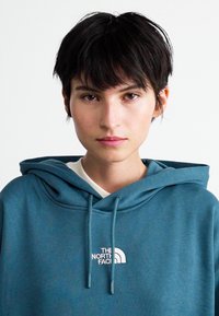 Jeune personne avec les cheveux courts et foncés portant un sweat à capuche bleu de The North Face, faisant face à l'avant sur un fond clair uniforme.