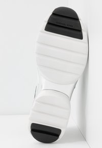 Semelle de chaussure blanche avec des zones d'adhérence noires au talon et aux orteils, avec le nom de la marque Michael Kors sur la partie supérieure noire.