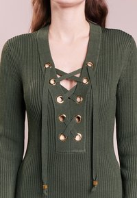 Top in maglia a costine verde oliva con scollo a V profondo, dettagli con occhielli dorati e laccetti incrociati al collo.