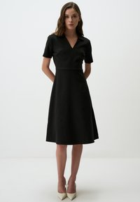 Schwarzes, kurzärmliges Kleid mit einem V-Ausschnitt, tailliertem Oberteil und ausgestellt Rock mit vertikalem Ripptextur, kombiniert mit spitzen beigen Absätzen.
