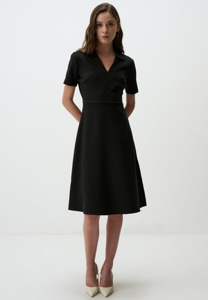 Schwarzes, kurzärmliges Kleid mit einem V-Ausschnitt, tailliertem Oberteil und ausgestellt Rock mit vertikalem Ripptextur, kombiniert mit spitzen beigen Absätzen.