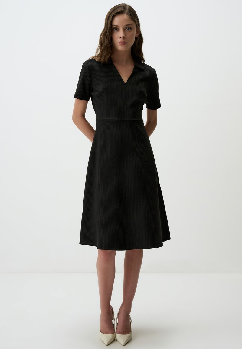 Schwarzes, kurzärmliges Kleid mit einem V-Ausschnitt, tailliertem Oberteil und ausgestellt Rock mit vertikalem Ripptextur, kombiniert mit spitzen beigen Absätzen.