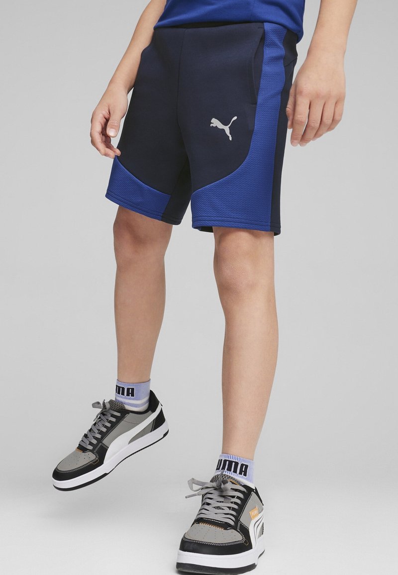 Personne portant un short Puma noir et bleu avec le logo Puma, des baskets grises et noires, et des chaussettes blanches Puma sur un fond gris.