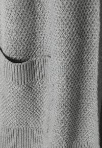 Pull en maille gris avec un motif texturé, bord-côte en bas et poche avant. Matière douce au toucher, offrant une apparence chaleureuse.