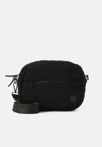 Kompakt svart fleeceminiatyrväska med framficka med dragkedja och justerbar axelrem mot enfärgad vit bakgrund.