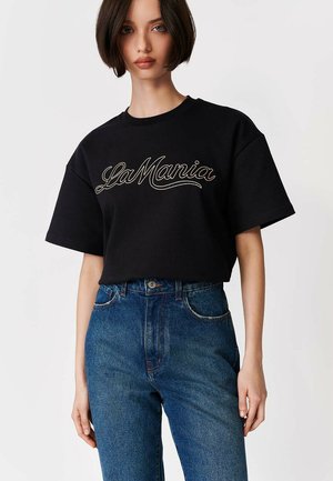 Kvinde med kort brunt hår iført en sort "La Mania" t-shirt og højtaljede blå jeans foran en ensfarvet hvid baggrund.