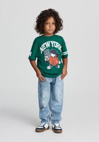 T-shirt verde oversize con stampa "NEW YORK" e grafica in stile cartoon, abbinata a jeans chiari strappati e sneakers blu navy/bianco.