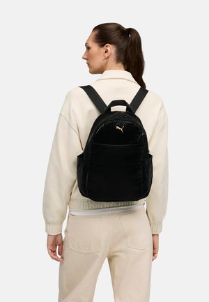 Mochila de terciopelo negro con logo dorado, con correas ajustables, dos bolsillos frontales y una forma redondeada. Combina con un suéter color crema.