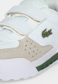 Baskets blanches en suède beige texturé, deux bandes Velcro et un logo crocodile vert sur le côté au-dessus d'une semelle verte et blanche.