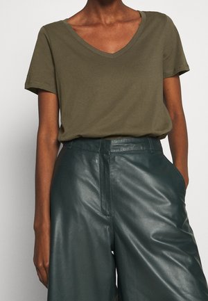 Persona que lleva una camiseta de manga corta de cuello en V de color verde oliva metida en unos pantalones de cuero verde oscuro de talle alto, con una mano en el bolsillo.