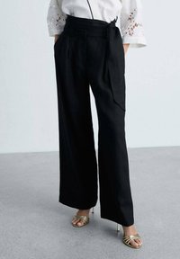 Pantaloni negri, cu talie înaltă și picioare largi, cu pliuri și bucle pentru curea. Asociati cu pantofi cu toc metalici, cu vârful deschis și bretele încrucișate.